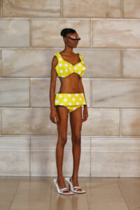 Model wearing a yellow polka-dot bikini with a ruffled bottom, standing against a beige stone wall.
