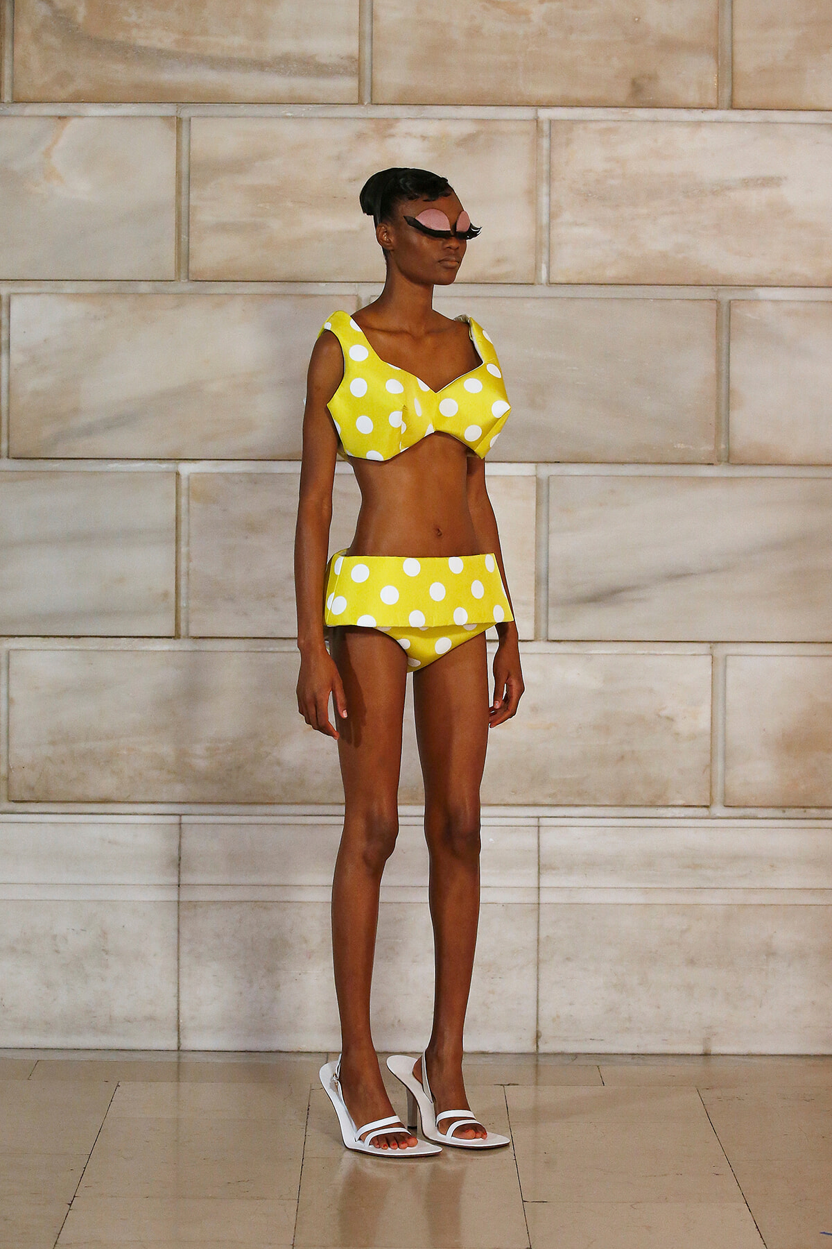 Model wearing a yellow polka-dot bikini with a ruffled bottom, standing against a beige stone wall.
