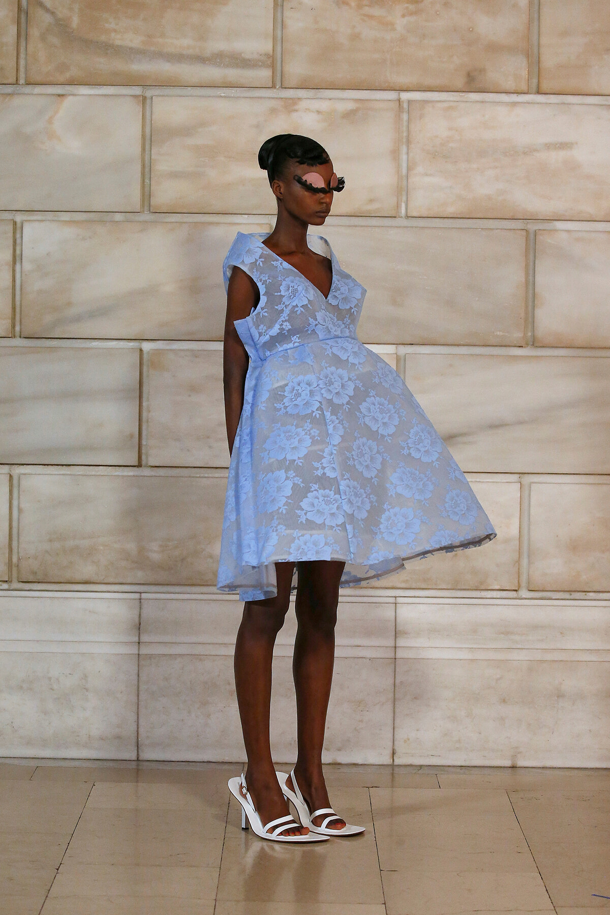 Model in a light blue floral lace dress, knee-length, with white sandals, standing against a beige stone wall.