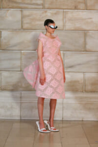 Model wearing a pink knee-length floral lace dress with cape-like sleeves, white heels, and large sunglasses, posing against a marble wall.