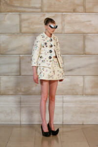 Runway model in a cream tweed jacket and mini skirt covered with colorful embellishments, wearing oversized sunglasses and black platform heels on a stone wall backdrop.