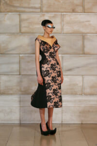 Model wearing a pink and black lace midi dress with beige collar and black side panels, standing against a stone wall.