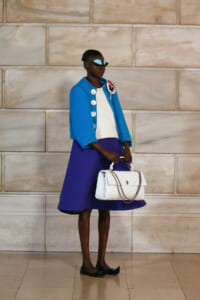 Woman in a cobalt blue jacket with large buttons over a white top and purple skirt, holding a white quilted handbag with a chain strap against a stone wall.