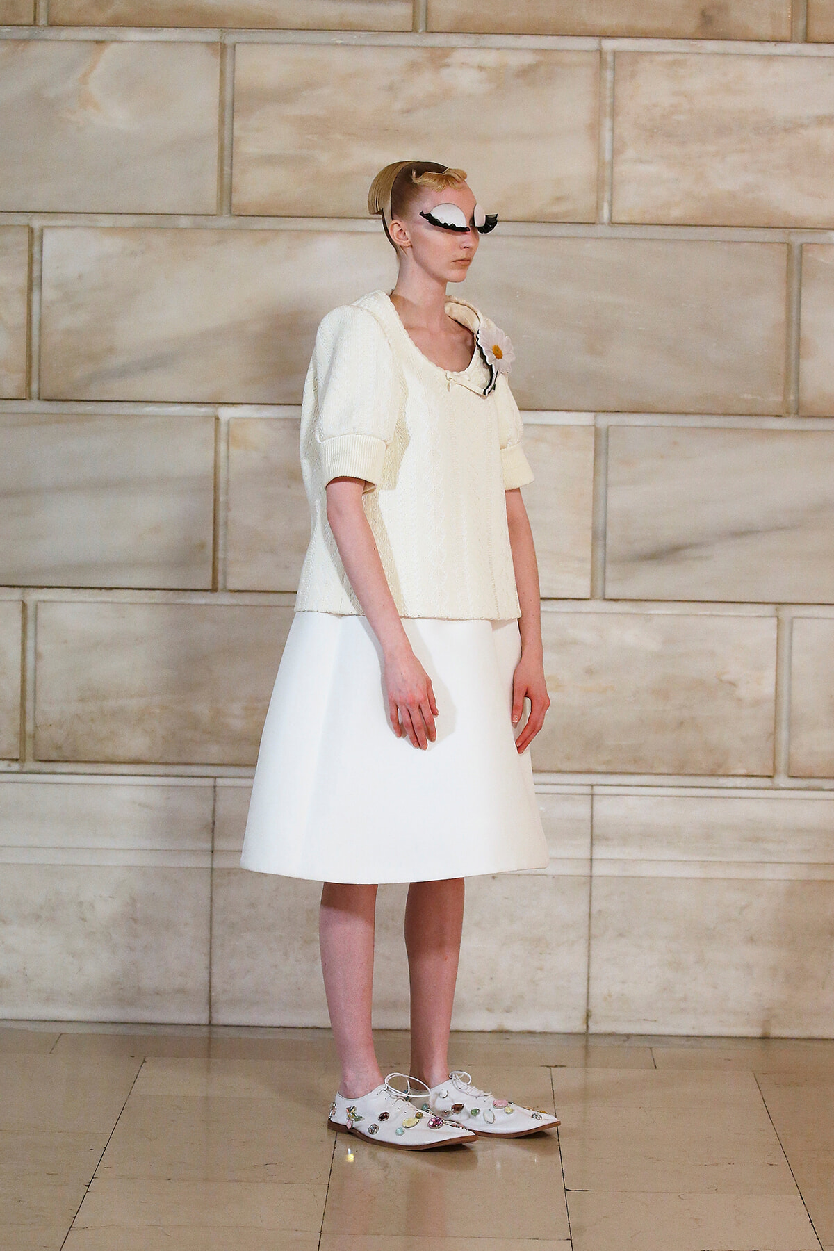 Model in a cream knitted top with puff sleeves and a white knee-length skirt, wearing white embellished shoes and a bold white eye mask with black lashes, posed against a beige stone wall.
