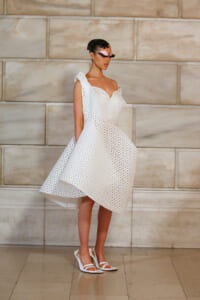 Model in a white, knee-length, geometric-pattern dress with an asymmetric neckline and cap sleeve, standing against a stone wall, wearing a pink masquerade mask and white heels.