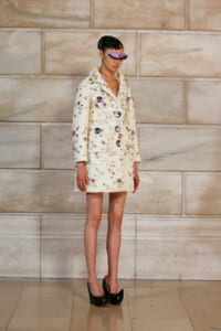 Model on runway wearing a cream, gem-covered coat with a lavender mask and black platform heels.