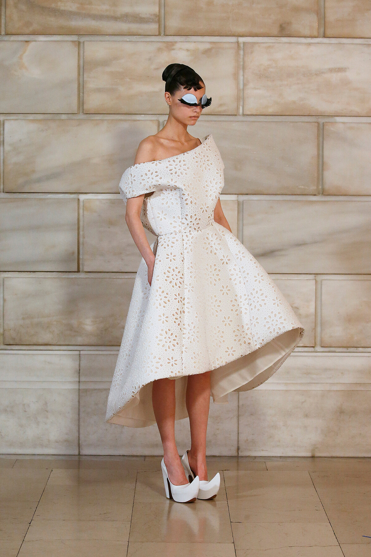 Model in an off-the-shoulder white eyelet dress with a high-low hem, bold white sunglasses, and white platform heels.