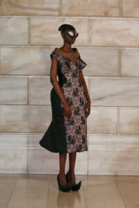 Model wearing a pink and black lace midi dress with off-the-shoulder neckline and black side panels, posing against a stone wall runway backdrop with black heels and pink sunglasses.