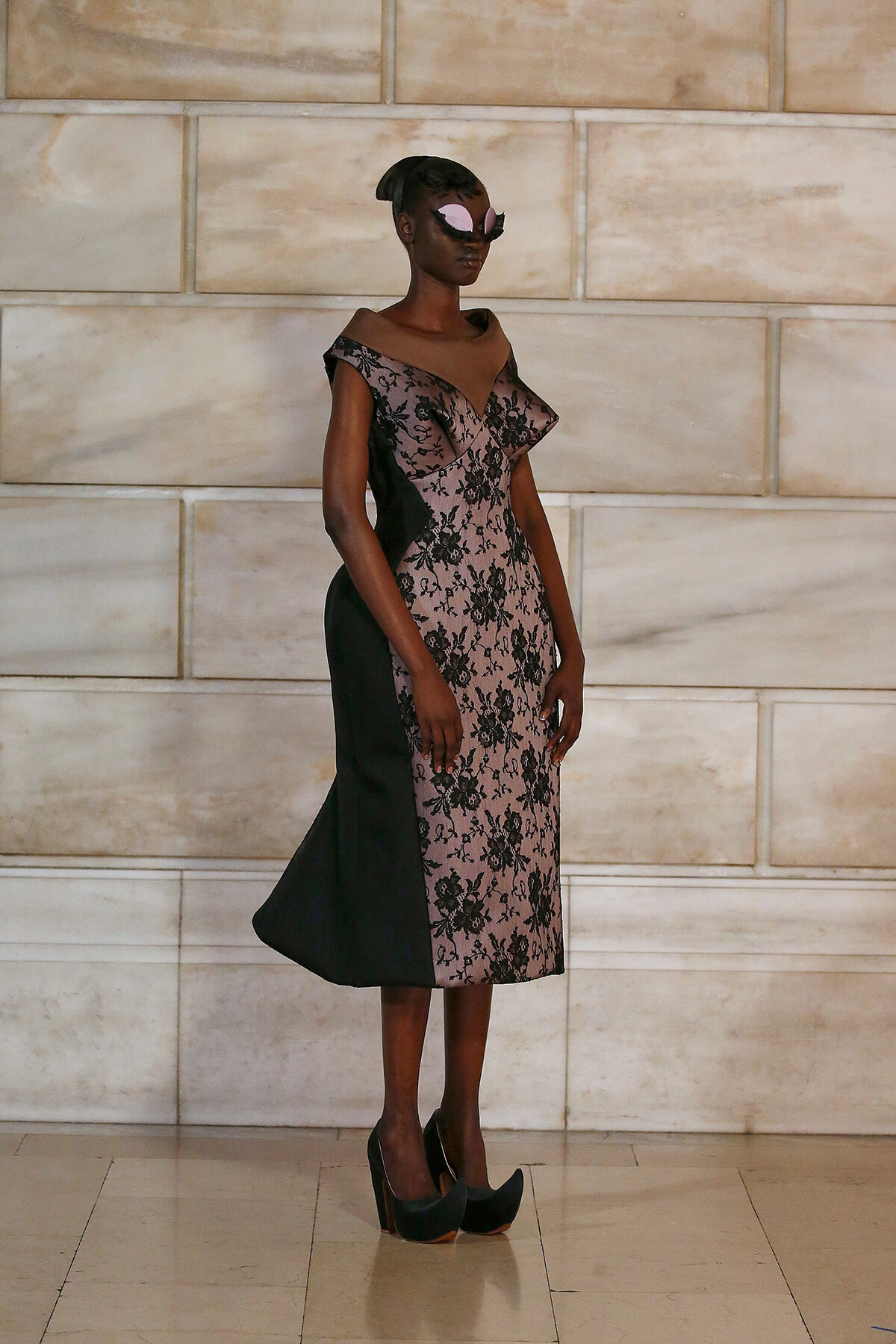Model wearing a pink and black lace midi dress with off-the-shoulder neckline and black side panels, posing against a stone wall runway backdrop with black heels and pink sunglasses.