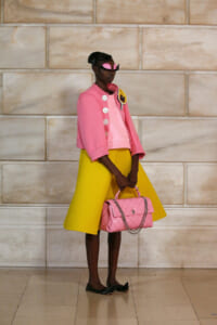 Model in a pink cropped jacket with white buttons, bright yellow skirt, pink quilted handbag, and pink mask, posed against a stone wall.