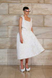 Model in a white sleeveless A-line dress with floral cutout pattern, paired with white platform heels, standing against a stone wall.