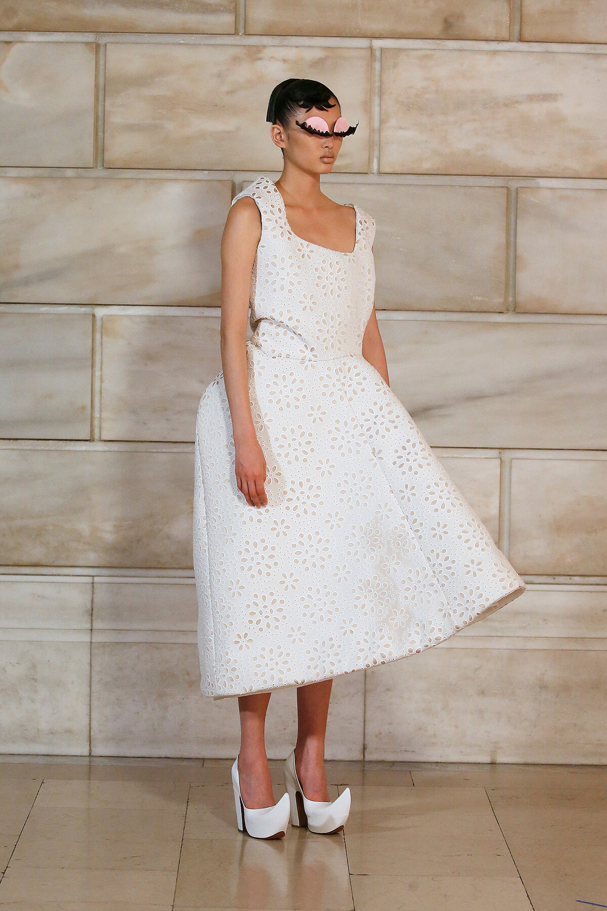 Model in a white sleeveless A-line dress with floral cutout pattern, paired with white platform heels, standing against a stone wall.