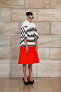Model wearing a white top with black stripes and a bright orange A-line skirt, black platform heels, and teal sunglasses, posing against a beige stone wall.