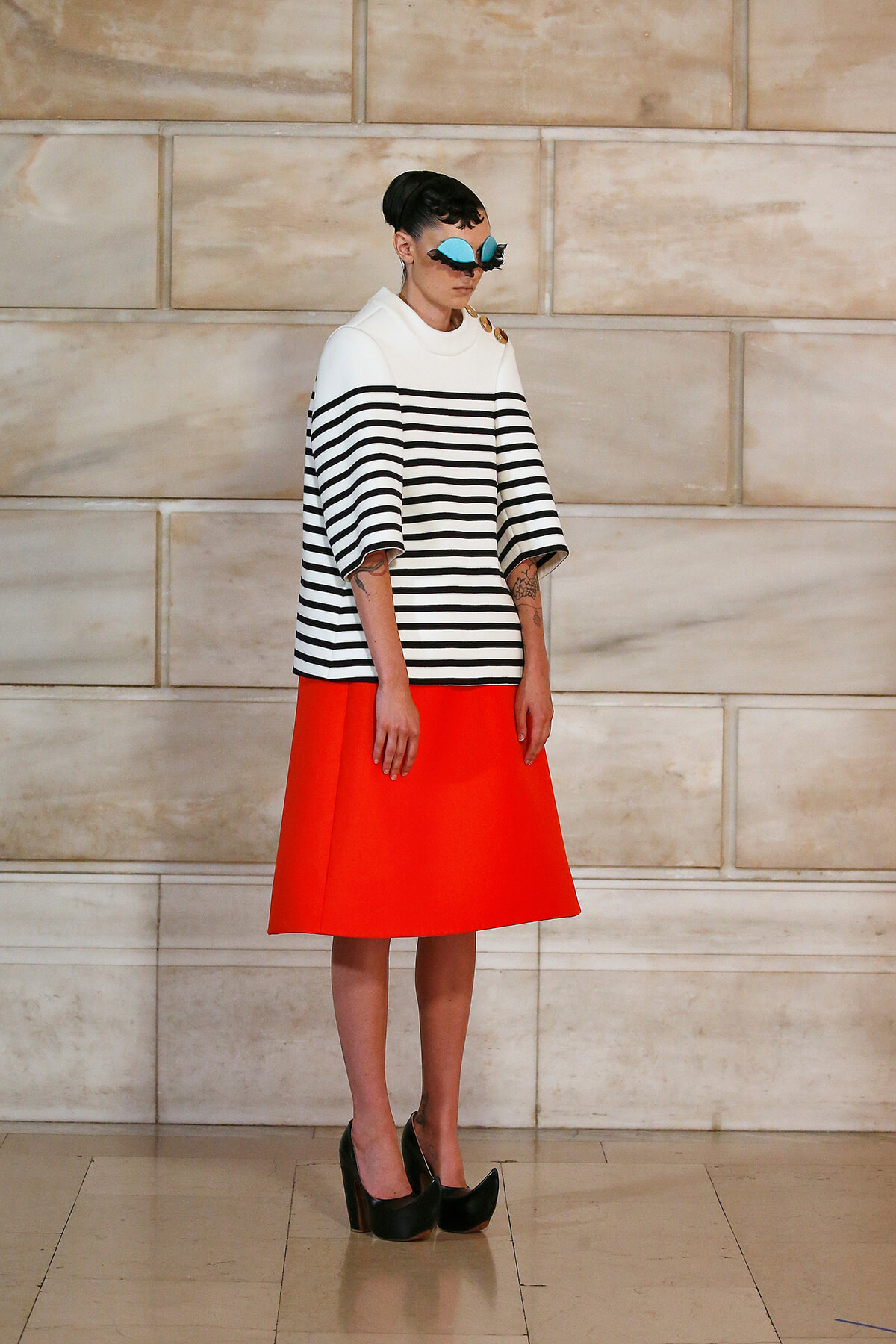 Model wearing a white top with black stripes and a bright orange A-line skirt, black platform heels, and teal sunglasses, posing against a beige stone wall.