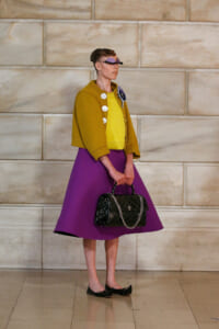Model in a mustard cropped jacket over a yellow top with a purple full skirt, carrying a black quilted handbag with a chain, and wearing sunglasses against a stone wall.