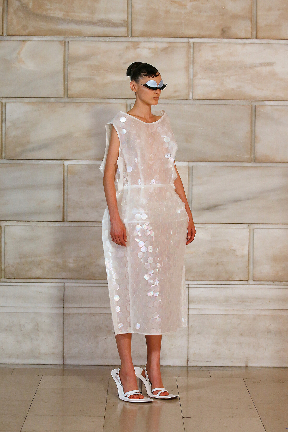 Model in a white sequined dress with large circular sequins, wearing a black-and-white mask, standing against a light stone wall.