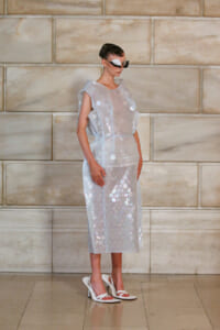 Model wearing a sheer, silver midi dress covered in large circular sequins, white heeled sandals, and a futuristic white mask against a stone wall backdrop.