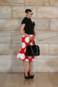 Model in a black top and bold red polka-dot skirt poses against a beige stone wall, holding a black handbag with chain.
