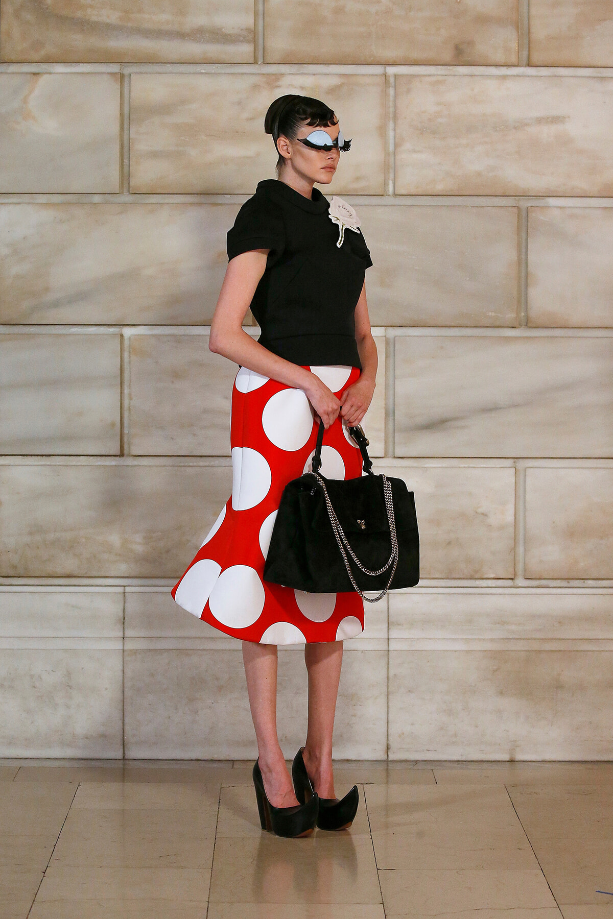Model in a black top and bold red polka-dot skirt poses against a beige stone wall, holding a black handbag with chain.
