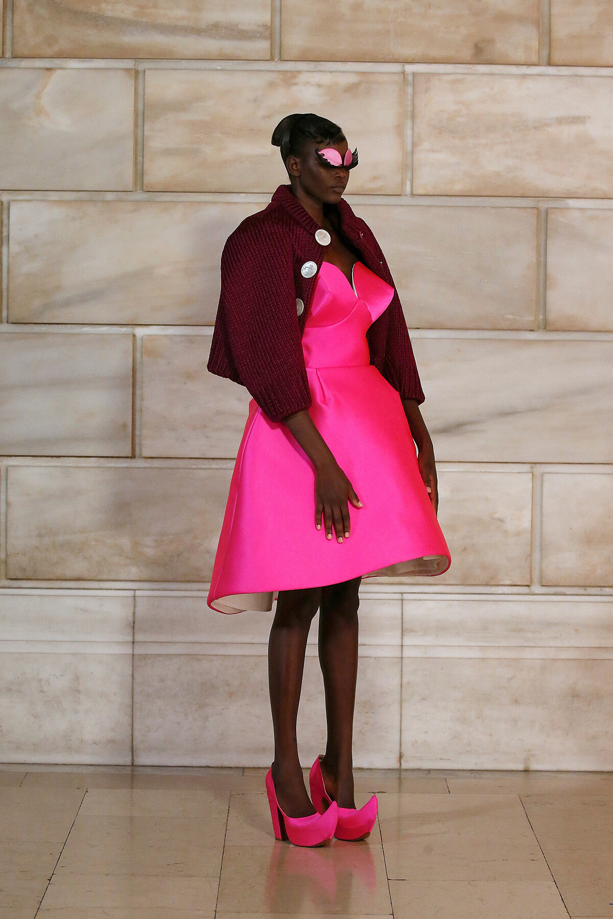Model wearing a bright pink dress with a burgundy cardigan and matching pink heels, posing against a light stone wall with a pink mask accessory.