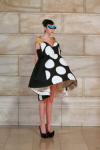 Model posing in a sculptural black dress with large white polka dots, yellow collar, and blue oversized sunglasses against a stone wall.