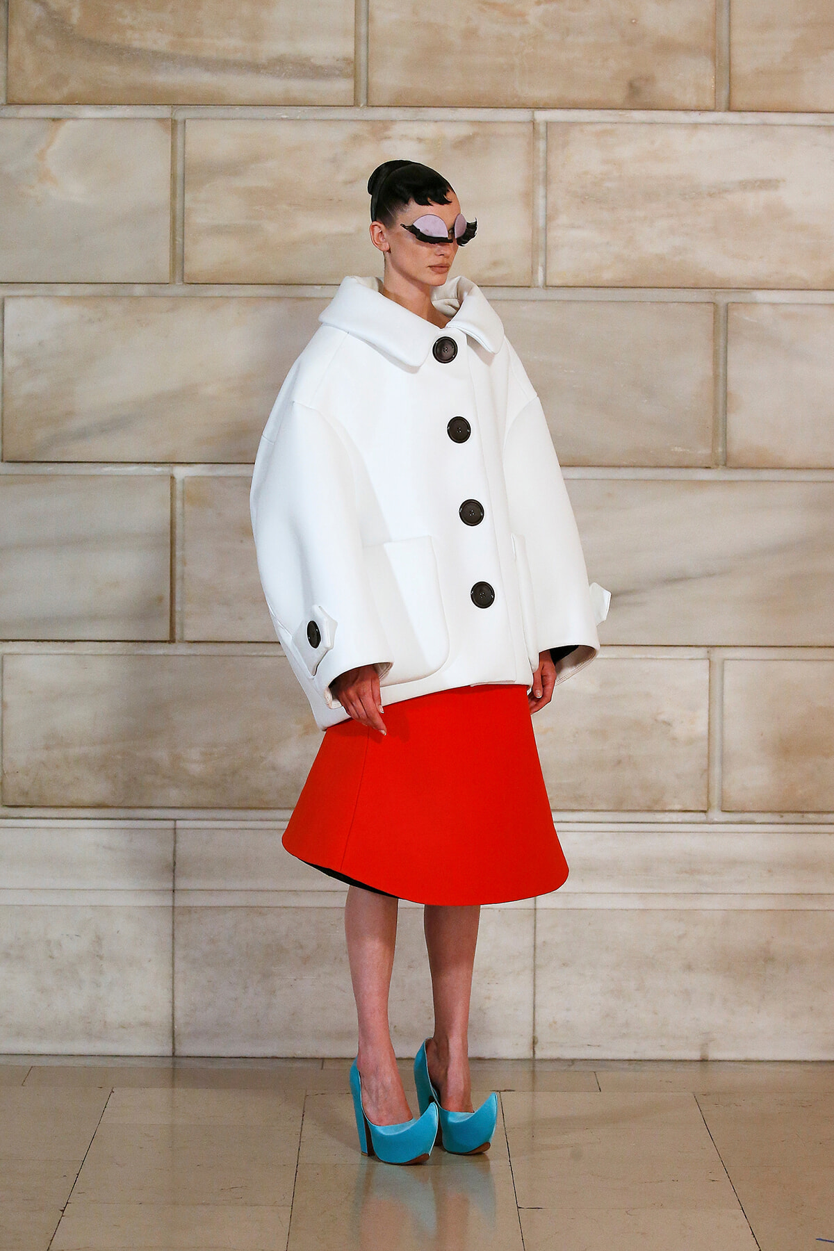 Model in an oversized white coat with large black buttons, a bright red A-line skirt, and turquoise platform heels on a runway set against a stone wall.