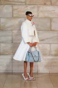 Model in an all-white high-fashion outfit with oversized sleeves, pink rose brooch, and light blue handbag with a silver chain, standing against a stone wall.