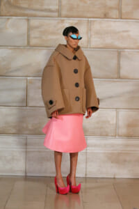 Model in an oversized camel coat with large black buttons, paired with a pink knee-length skirt and bright pink heels, standing against a stone wall.