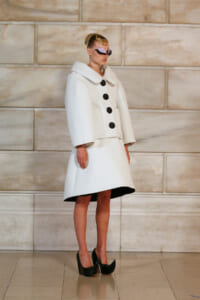 Model in an oversized white coat with a large collar and five black buttons, paired with a white A-line skirt and black heels, posing against a stone wall.