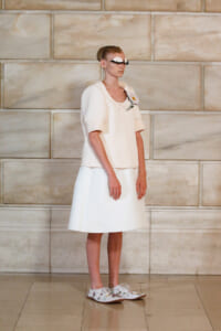 Model in an all-white outfit on a runway: cable-knit short-sleeve sweater over a knee-length skirt, flower brooch, playful goggles, and embroidered loafers.