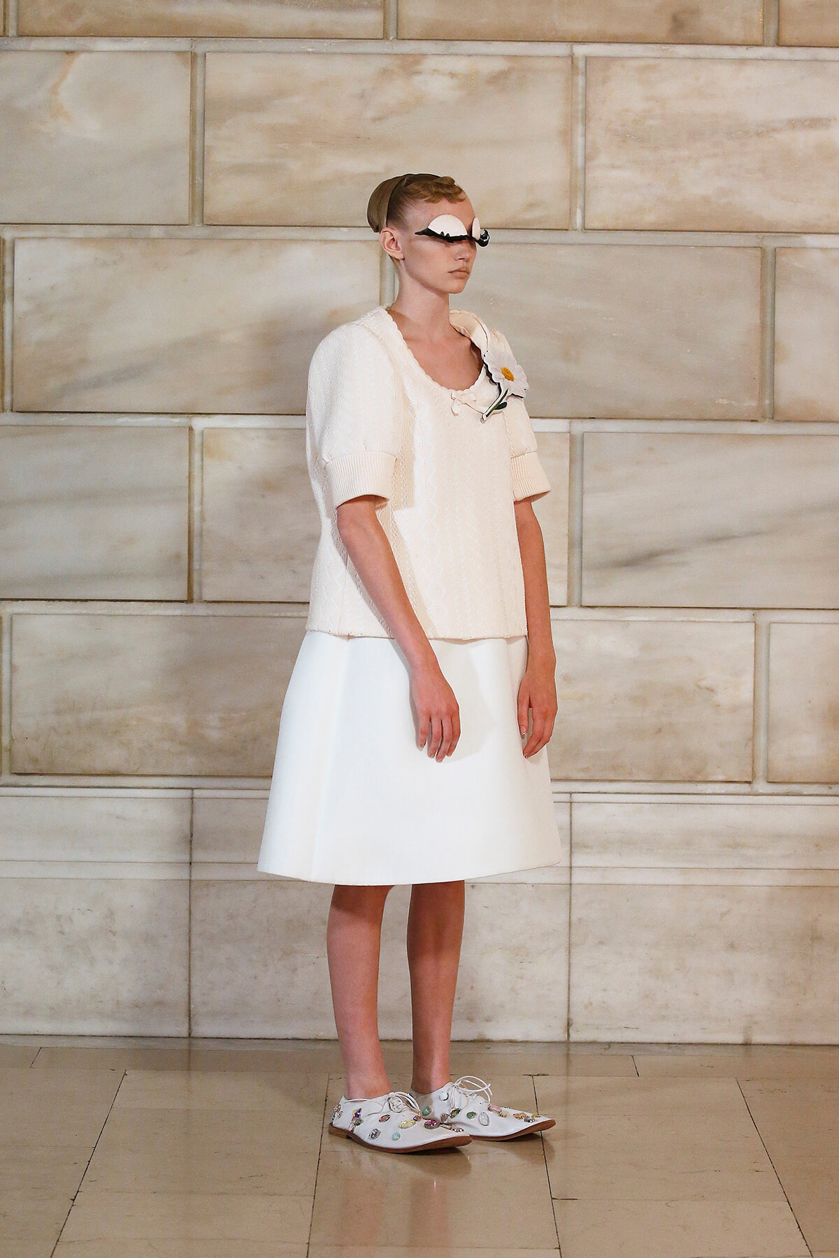 Model in an all-white outfit on a runway: cable-knit short-sleeve sweater over a knee-length skirt, flower brooch, playful goggles, and embroidered loafers.