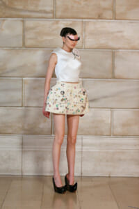Model in a white sleeveless top and a cream mini skirt covered in colorful gems, with black platform heels, posing on a runway against a stone wall.