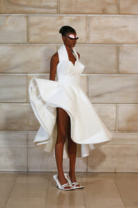 Model in a white sculptural gown with a voluminous, swirling skirt and pink masquerade mask against a stone wall backdrop.