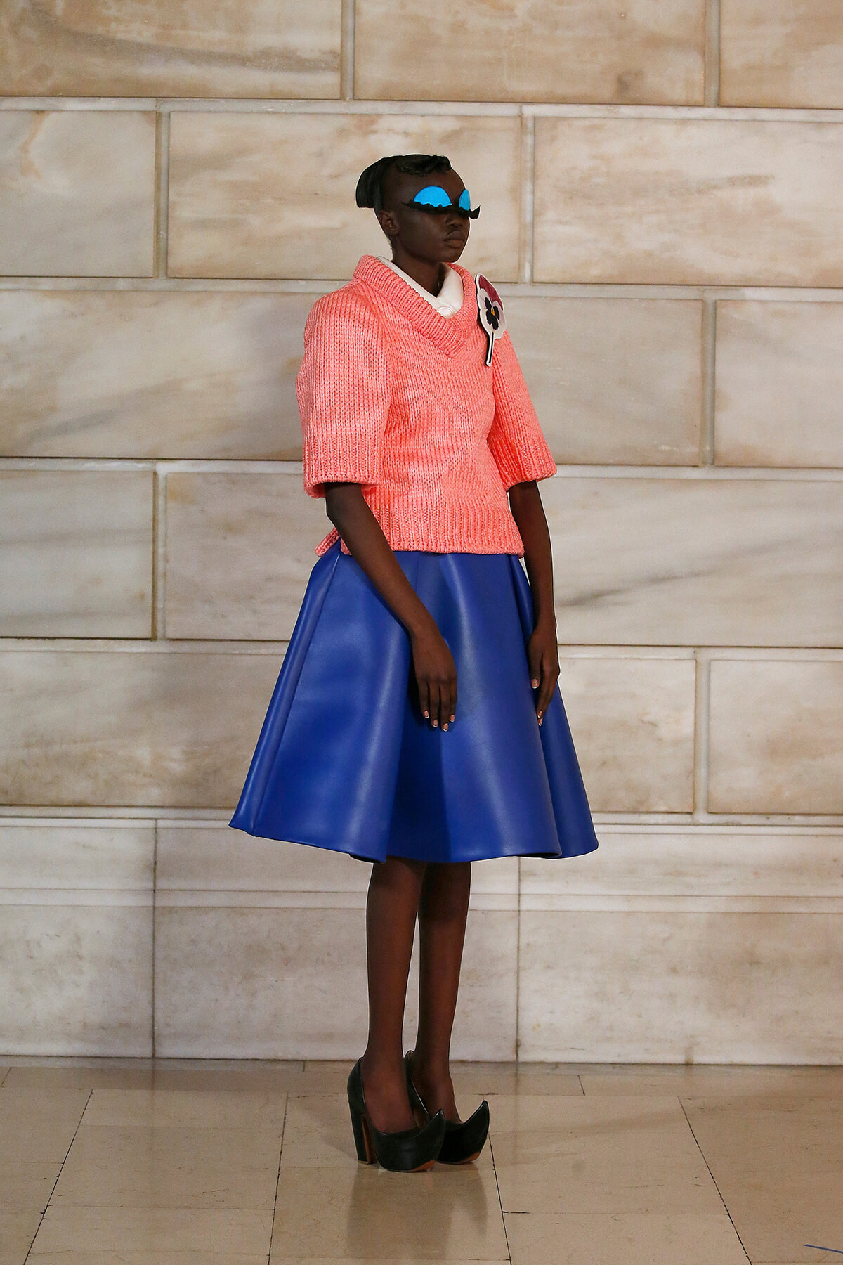 Model standing against a stone wall in a chunky pink knit sweater with a cobalt blue A-line skirt and black high heels, wearing blue sunglasses and a floral brooch.