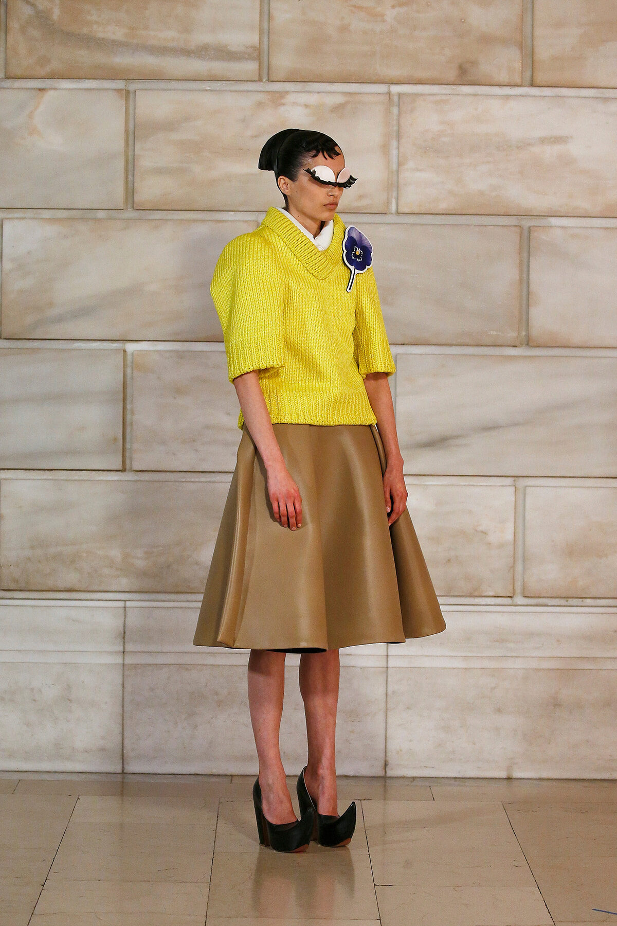 Model wearing a bright yellow knit sweater with a beige A-line skirt and black high heels, standing against a stone wall. She wears a decorative mask and a purple flower brooch on the sweater.