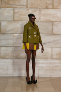 Model wearing an olive green military-style coat with large silver buttons, paired with a color-blocked yellow and brown skirt, and black high heels, standing against a light stone wall.