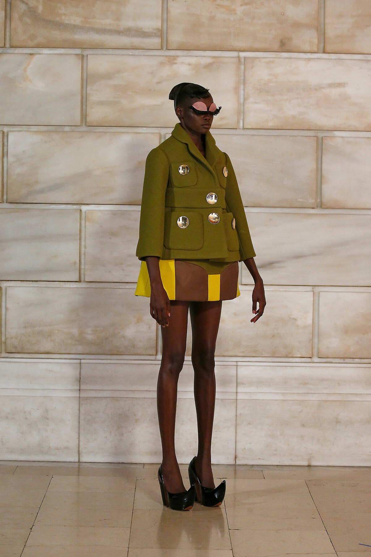 Model wearing an olive green military-style coat with large silver buttons, paired with a color-blocked yellow and brown skirt, and black high heels, standing against a light stone wall.