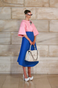 Model wearing a pink oversized top with a blue midi skirt, white handbag, and white platform heels against a stone wall.