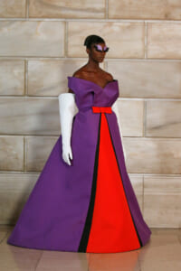 Model wearing a dramatic purple evening gown with a red center panel and black trim, paired with white opera gloves and a purple feathered mask.