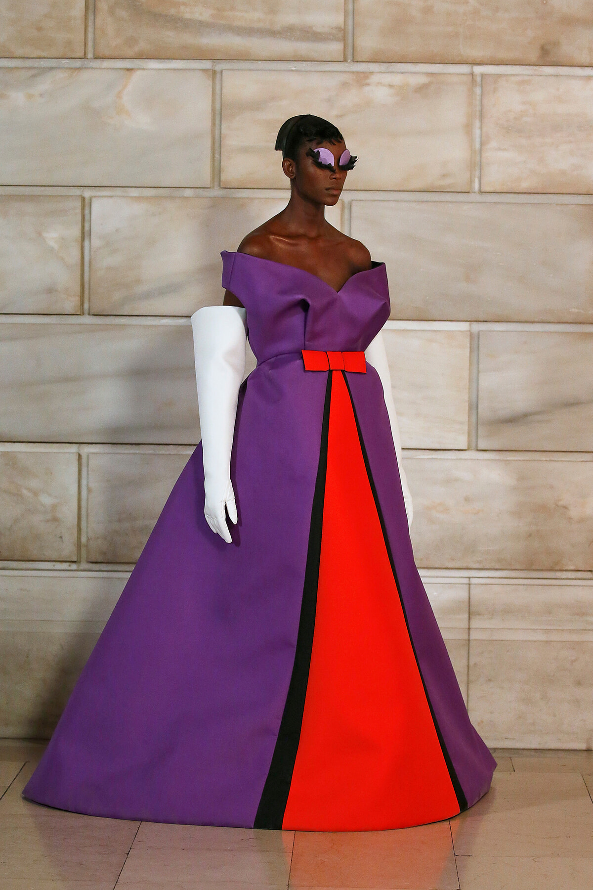 Model wearing a dramatic purple evening gown with a red center panel and black trim, paired with white opera gloves and a purple feathered mask.