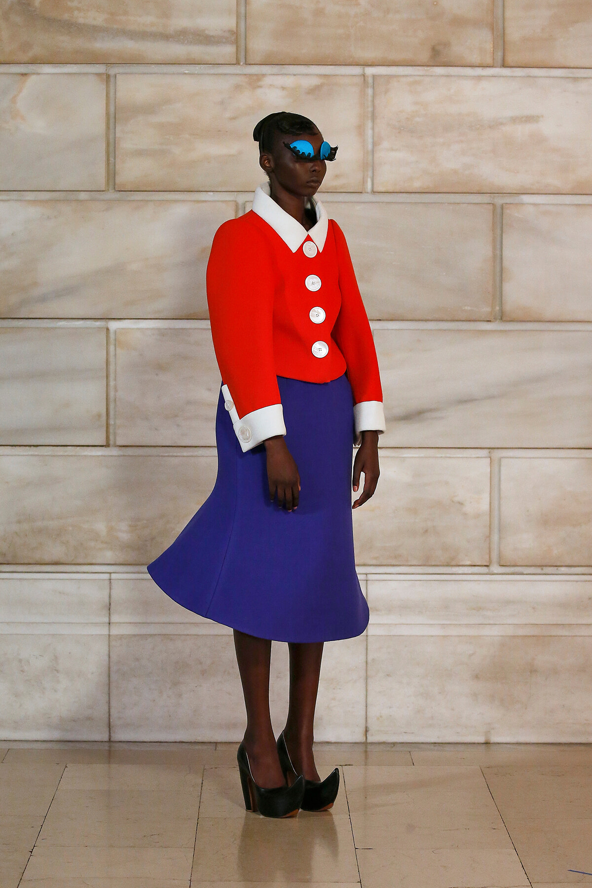Model wearing a bright red structured jacket with white collar and cuffs, a cobalt blue A-line skirt, blue sunglasses, and black platform heels against a stone wall.