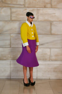 Full-length model in a bright yellow jacket with white collar and cuffs, a purple midi skirt, purple mask, and black high heels against a stone wall.