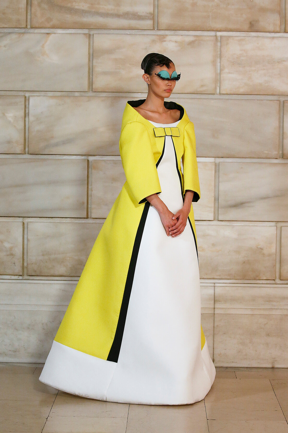 Model in a bright yellow cape-style coat over a white gown with black trim and a yellow bow, standing against a stone wall, wearing teal masquerade mask and poised posture.