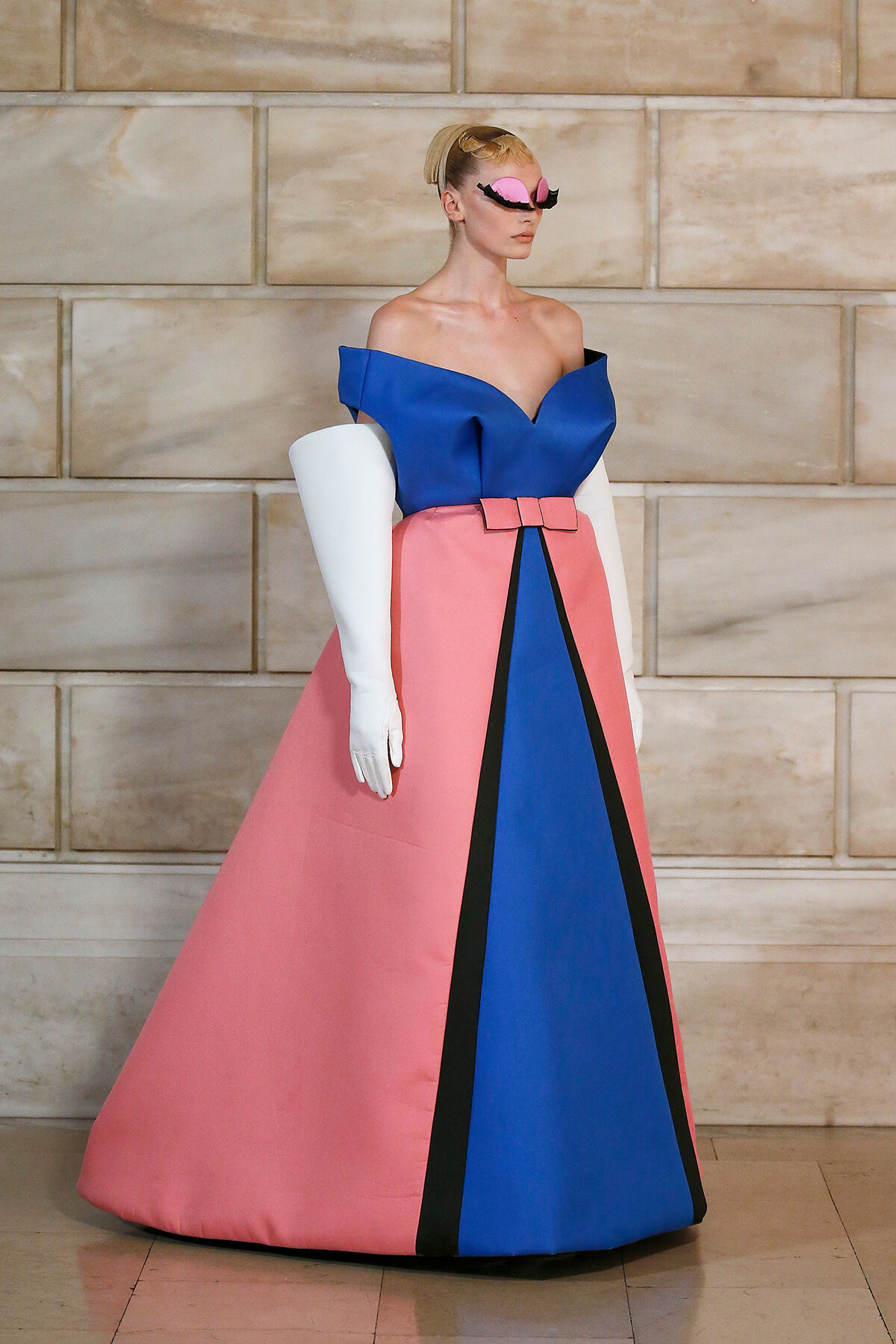 Model in a color-block gown: blue off-the-shoulder bodice, pink skirt with black and blue panels, and white opera gloves; pink mask-like sunglasses.