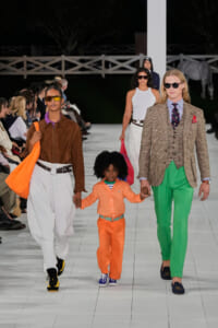 Runway scene: a man in a plaid blazer and bright green pants walks beside a woman in a brown suede jacket and white pants; they hold a child’s hands as they walk.