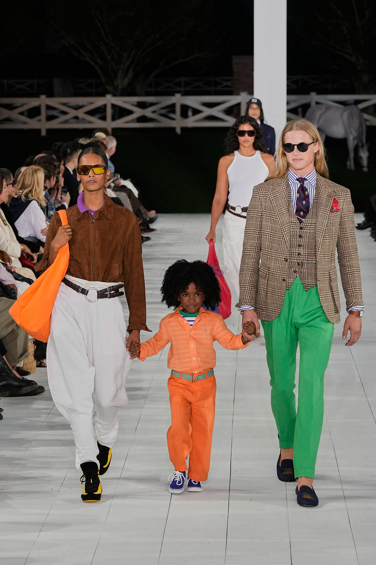 Runway scene: a man in a plaid blazer and bright green pants walks beside a woman in a brown suede jacket and white pants; they hold a child’s hands as they walk.