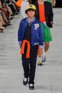 Young boy model walking a fashion runway in a blue varsity jacket, striped shirt, pinstripe pants, yellow Yankees cap, orange sweater tied around waist, navy sneakers.