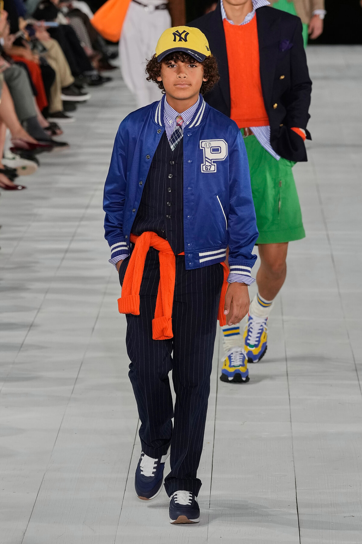 Young boy model walking a fashion runway in a blue varsity jacket, striped shirt, pinstripe pants, yellow Yankees cap, orange sweater tied around waist, navy sneakers.