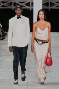 Two models walk the runway: a man in a white double‑breasted suit with a bow tie and plaid pants, wearing a navy cap; a woman in a cream satin slip dress with a wide belt and a red handbag.