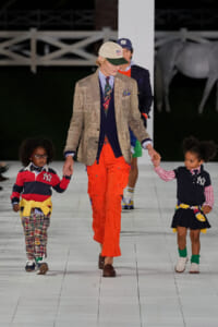 Adult model walks a white runway holding hands with two children in colorful outfits; beige plaid blazer, navy vest, striped shirt, and bright orange pants.